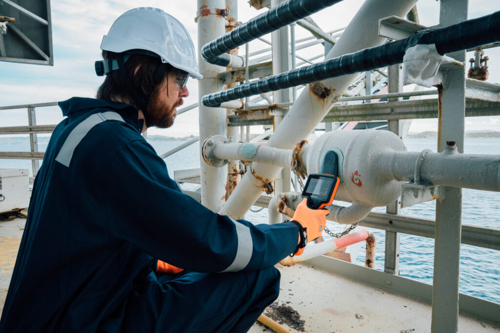 A worker in protective gear, including helmet and gloves, uses a WAND-HDC device to inspect pipes on an offshore platform, with the sea in the background.