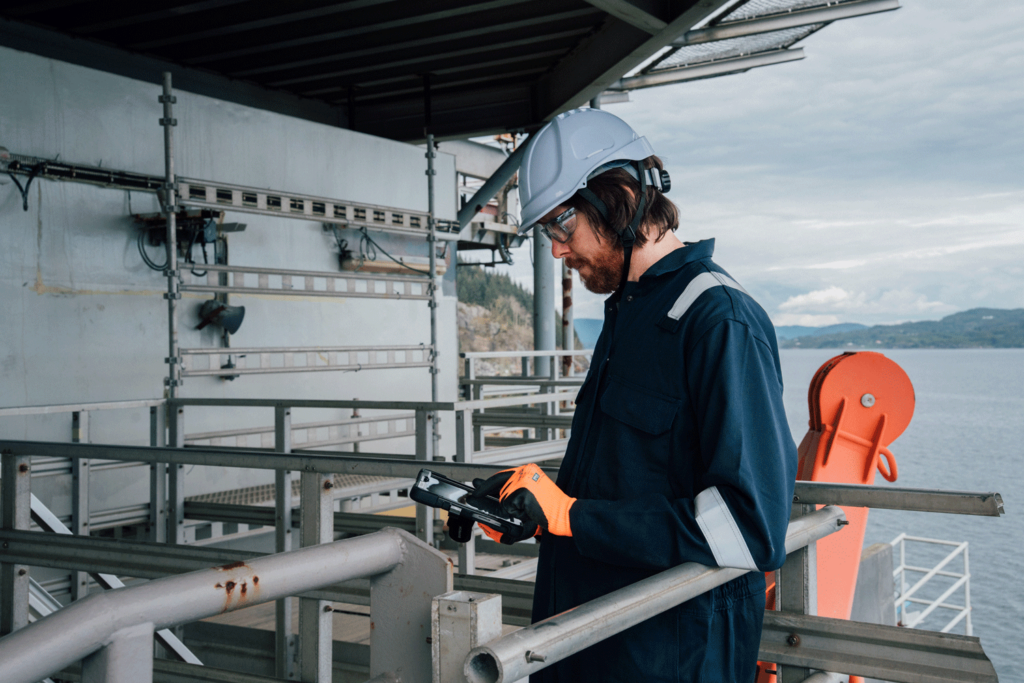 A worker in safety gear and a hard hat stands on an industrial platform overlooking water, using a tablet device.