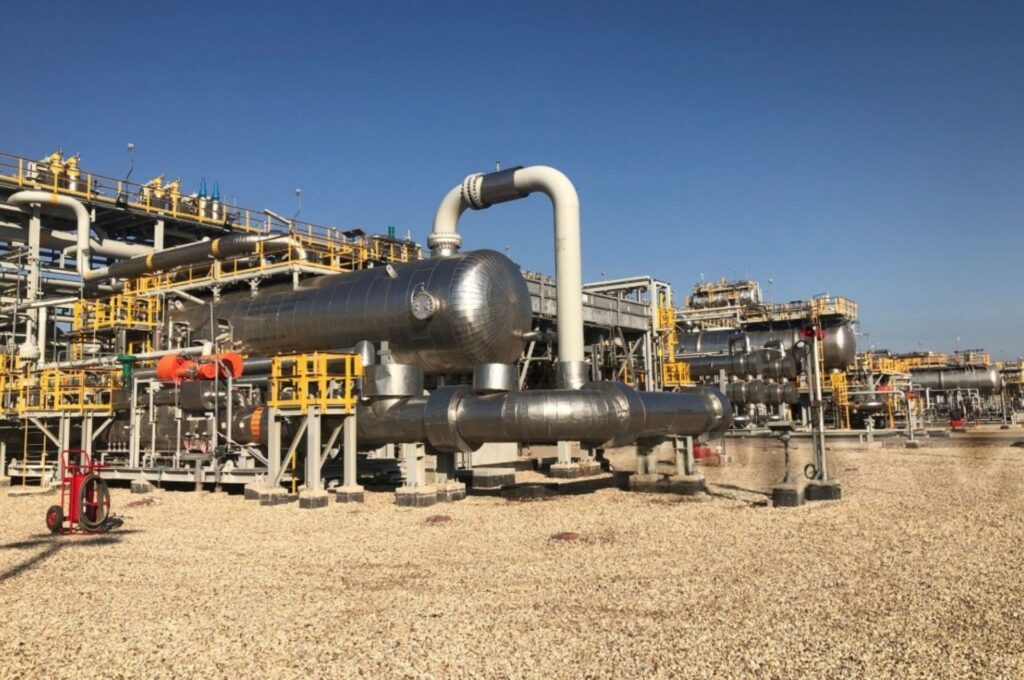 Industrial plant with large silver pipelines and storage tanks under a clear blue sky. Yellow railings add contrast; a sense of scale and industry.